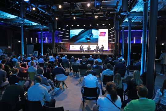 A congress in a modern remodelled industrial hall with atmospheric blue lighting. A panel discussion is taking place on the stage with four people sitting on chairs. Numerous spectators in business attire sit in the auditorium and follow the discussion attentively.