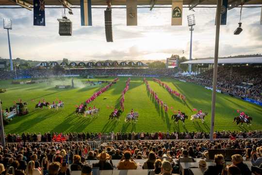 Blick ins ausverkaufte Hauptstadion während der Eröffnungsfeier. Auf dem Rasen befinden sich zahlreiche Tänzer in vier Reihen, die ganz in Pink gekleidet sind, inspiriert vom Barbie Thema und passend zum Partnerland USA. Zum Schaubild gehören außerdem eine Gruppe von pink gekleideten Reitern, die in Zweierpaaren auf dem Weg zum Ausritt sind und alle einen pinken Luftballon an einer Schnur festhalten.