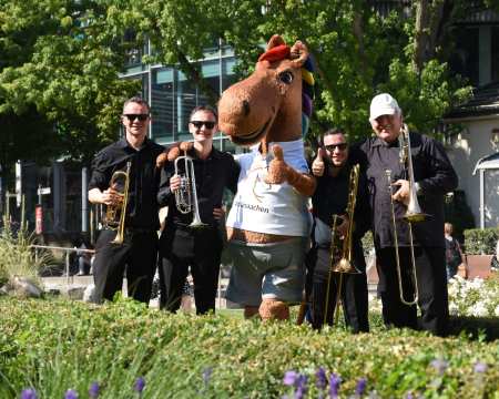 A group of four musicians dressed in black stand in the greenery with trumpets and trombones next to a large, cheerful horse mascot wearing a white T-shirt labelled #chioaachen.
