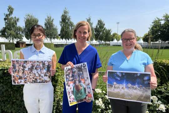 Drei Personen stehen nebeneinander im Freien vor blühenden weißen Rosen und halten jeweils ein großes Farbfoto vor sich: Die linke Person präsentiert ein Bild mit einer jubelnden Menschenmenge, die mittlere Person hält ein Foto mit einem Reiter auf einem Pferd, die rechte Person zeigt ein Bild mit einer Pferdeszene im Wasser. Im Hintergrund sind grüne Wiesen, Bäume und ein Teil einer weißen Tribüne zu sehen.