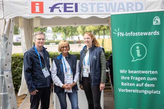 Drei Personen stehen vor einem weißen Zelt mit der Aufschrift „FEI Steward“. Sie tragen blaue Jacken oder Westen mit FN-Logo und Namensschildern um den Hals. Rechts im Bild steht ein grünes Roll-up mit der Aufschrift „FN-Infostewards – Wir beantworten Ihre Fragen zum Reiten auf dem Vorbereitungsplatz“. Im Hintergrund sind Bäume und ein Turnierplatz zu sehen.