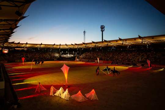 Das Dressurstadion bei Abenddämmerung, der Sandplatz ist in farbiges Licht getaucht. Vor vollen Tribünen findet eine Show statt: Tänzerinnen in leuchtenden Kostümen bewegen sich am Rand der Arena, während mehrere Reiter mit beleuchteten Pferden in der Mitte auftreten.