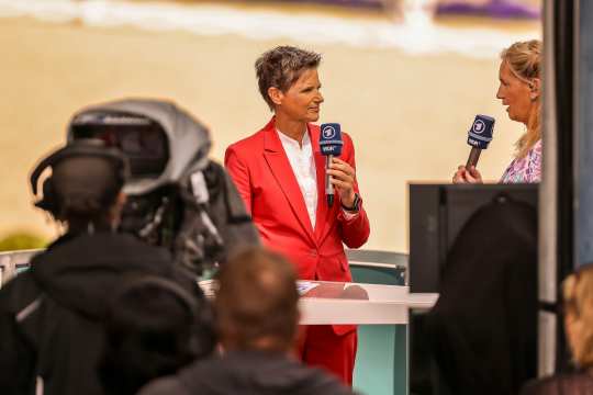 Zwei Frauen stehen in einem Fernsehstudio an einem Tisch und halten Mikrofone mit dem Logo von WDR und ARD. Eine Person trägt einen roten Anzug, die andere ein gemustertes Oberteil. Im Vordergrund ist ein Kamerateam zu sehen, im Hintergrund lässt sich unscharf das Dressurstadion erahnen.