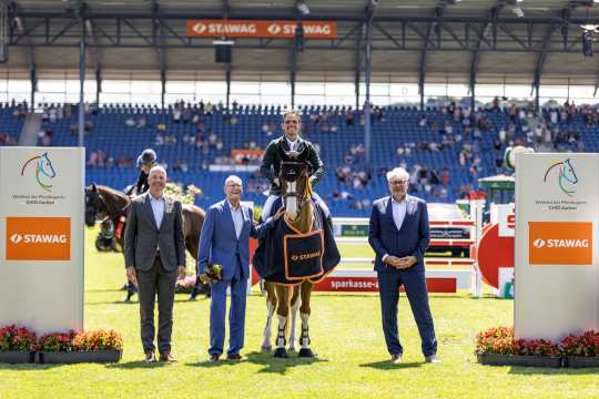 The photo shows a rider in a green jacket on a brown horse with a white blaze and four white legs. The horse is wearing a dark blue winner's blanket with orange edging and the STAWAG logo. To the left and right of the horse are three men in suits, one holding a bouquet of flowers. In the background, blue stands with spectators, obstacles and large signs with the CHIO Aachen logo and STAWAG lettering can be seen. The scene takes place on the sunny grass arena.