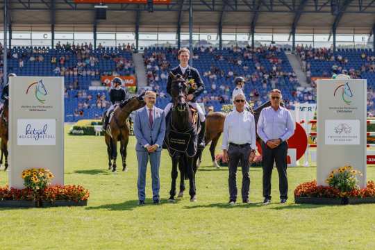 A rider in a dark jacket sits on a dark brown horse with a black winner's blanket bearing the words ‘Deutschlands U25 Springpokal’ in gold lettering. The rider is holding a bouquet of flowers in his hand. To the left and right of the horse are three men, one in a blue suit and the others in light blue shirts. In the background are more riders, spectator stands with lots of people and two large signs with the CHIO Aachen logo and sponsor logos. In front of the signs are floral decorations and colourful flowers.