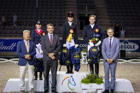 Siegerehrung bei einem Reitturnier im Stadion: Drei Reiterinnen und Reiter in Turnierkleidung und Helmen stehen auf dem Podest, umgeben von vier offiziellen Personen in Anzügen. Die Reiterinnen und Reiter tragen Schleifen, halten Blumensträuße und Siegerprämien in den Händen. Im Hintergrund sind Tribünen und Werbebanden zu sehen.