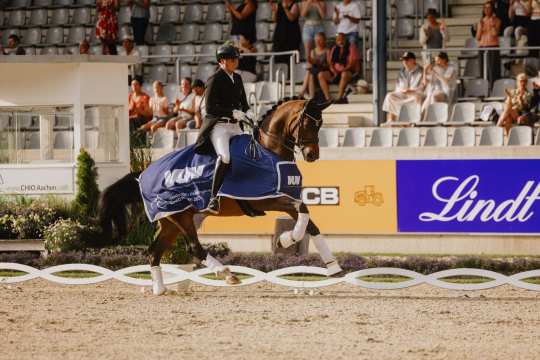 Ein Reiter in Turnierkleidung reitet ein dunkelbraunes Pferd, das eine Siegerschabracke mit der Aufschrift "Weltfest des Pferdesports CHIO Aachen 2025" trägt. Im Hintergrund sitzen und stehen Zuschauer auf den Tribünen, während Werbebanden mit Sponsorenwerbung zu sehen sind.