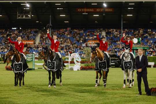 Four riders in red jackets sit next to each other on their horses, which are wearing black winners' blankets with the Mercedes logo. Three of them raise their riding helmets in salute, one person waves. In the background, many spectators can be seen in the stands of a large, floodlit stadium. On the right, a person in a suit stands on the pitch.