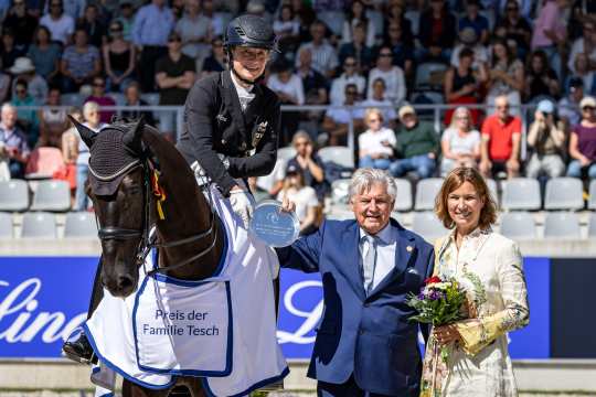 A dressage rider rides a dark brown horse in a dressage competition. A large display board with the name ‘Isabell Werth’ and spectators in the stands can be seen in the background. The rider is wearing a black jacket, white breeches and a helmet. The scene takes place in a stadium with floral decorations and a dressage arena.