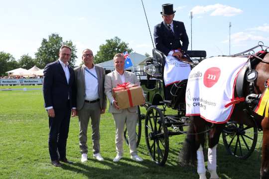 Three men in suits stand in a meadow next to a festively decorated carriage. One of the men is holding a gift with a red ribbon in his hands. A driver in a tailcoat and top hat is sitting on the carriage. The horse is decorated with a white blanket with logos and a German ribbon. Tents, trees and a blue sky can be seen in the background.