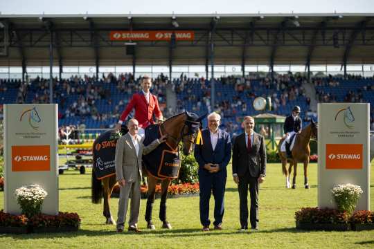 A rider in a red jacket sits on a decorated horse with a winner's blanket and ribbon. Three elegantly dressed men are standing next to the horse. A full grandstand, other horses and riders as well as two large boards with the CHIO Aachen logo and the sponsor STAWAG can be seen in the background. Flower arrangements decorate the foreground.