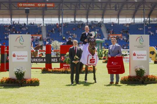 Award ceremony in the Sparkasse-Youngsters-Cup: A rider sits on his horse, which is wearing a winner's blanket with the Sparkasse logo. Two people stand to his left and right, one holding flowers, the other a red bag. Obstacles, advertising boards and spectators in the stands can be seen in the background.
