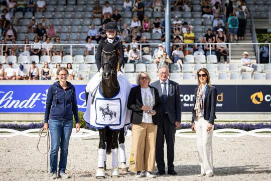 A dressage rider sits on a black horse wearing a white and blue winner's blanket. To the right of the horse are three elegantly dressed people and to the left a person in casual clothes with a win rope in his hand. One of the people is holding a large glass trophy. Spectators in the stands and advertising boards can be seen in the background.