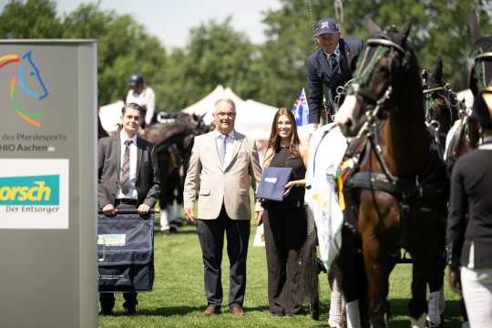 Several people and a team of horses are standing on a competition field at CHIO Aachen. On the left, there is a sign with the CHIO Aachen logo and the sponsor Horsch. A woman is holding a blue folder, a man in a suit is holding a horse rug, and another man stands in the center. In the background, more horses and participants can be seen.