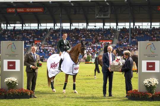 A rider in a green jacket sits on a horse with a white winner's blanket, on which the ‘Käfer’ logo and ‘World Equestrian Festival CHIO Aachen’ can be seen. Standing next to the horse are three men in suits, one holding a bouquet of flowers, another handing over a large gift basket. A large spectator stand and other horses can be seen in the background. To the left and right in the foreground are two large panels with the CHIO Aachen logo and the Käfer logo, with floral decorations in front of them.