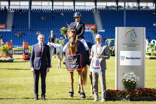 Three people and a horse are standing on a showground in the stadium. The person in the middle is sitting on a brown horse wearing a winner's blanket with the NetAachen logo. On the left is a person in a suit, on the right a person in a light-colored jacket with a NetAachen bag. Other riders, spectators in the stands and a sign with the CHIO Aachen and NetAachen logos can be seen in the background.
