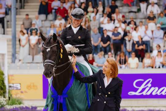 Eine Reiterin in Turnierkleidung sitzt auf einem dunkelbraunen Pferd mit einer blauen und grünen Siegerschabracke. Neben dem Pferd steht eine Frau im dunklen Blazer und legt die Hand auf den Hals des Pferdes. Im Hintergrund sind zahlreiche Zuschauer auf der Tribüne zu sehen.