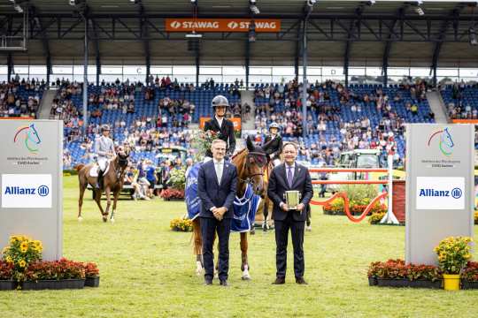 A rider in competition attire sits on a brown horse with a blue winner's saddle pad and holds a bouquet of flowers. Two men in suits stand in front of the horse, one holding a trophy. Other riders and numerous spectators in the stands can be seen in the background. To the left and right of the picture are large boards with the CHIO Aachen logo and the Allianz logo.