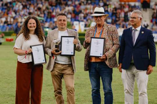 Vier Personen stehen auf einem Rasenplatz vor einer vollen Tribüne. Drei von ihnen halten gerahmte Urkunden in den Händen, eine Person zusätzlich eine Trophäe. Die Personen sind festlich gekleidet, eine trägt einen weißen Hut und ein buntes Sakko. Im Hintergrund sind viele Zuschauer auf der Tribüne zu sehen.