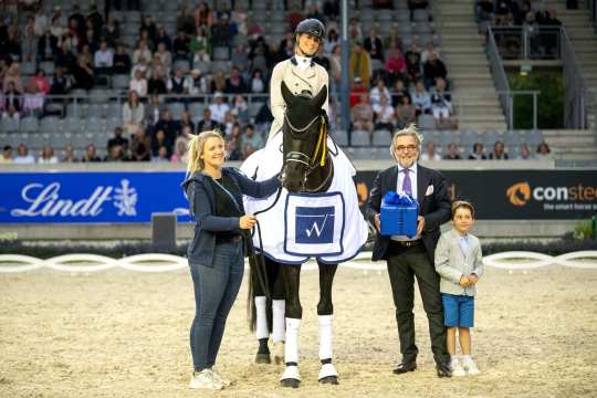 Eine Reiterin im hellen Reitoutfit sitzt auf einem geschmückten Pferd mit Siegerdecke in einer Arena. Neben dem Pferd stehen eine Frau, die das Pferd am Zügel hält, sowie ein Mann im Anzug mit einer blauen Geschenkbox und ein Junge in festlicher Kleidung. Im Hintergrund sind Tribünen mit zahlreichen Zuschauern zu sehen und Werbebanner an der Bande
