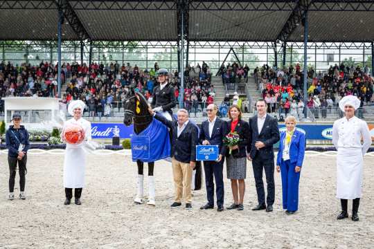 An award ceremony in an equestrian arena: a rider sits on a dark brown horse with a blue Lindt winner's blanket. Next to the horse are several people, including officials, a woman with a bouquet of flowers, a man with a Lindt bar and two people in white confectioners' uniforms with large chef's hats. In the background are stands filled with spectators.