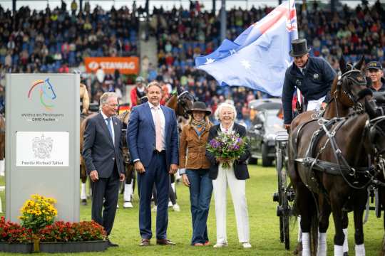 Prize-giving ceremony at the CHIO Aachen: In the foreground, several people are standing on the grass of the main stadium, including an elderly lady with a bouquet of flowers. On the right of the picture is a coachman with a top hat on a team of horses carrying an Australian flag. On the left is a sign with the CHIO Aachen logo and the name "“Familie Richard Talbot”. The full stands of the stadium can be seen in the background.