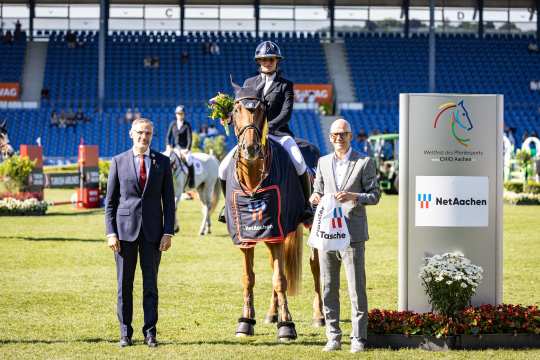 Drei Personen und ein Pferd stehen auf einem Turnierplatz im Stadion. Die Person in der Mitte sitzt auf einem braunen Pferd, das eine Siegerdecke mit dem Logo von NetAachen trägt. Links steht eine Person im Anzug, rechts eine Person im hellen Sakko mit einer NetAachen-Tasche. Im Hintergrund sind weitere Reiterinnen und Reiter, Zuschauer auf den Tribünen sowie ein Schild mit dem Logo des CHIO Aachen und NetAachen zu sehen.