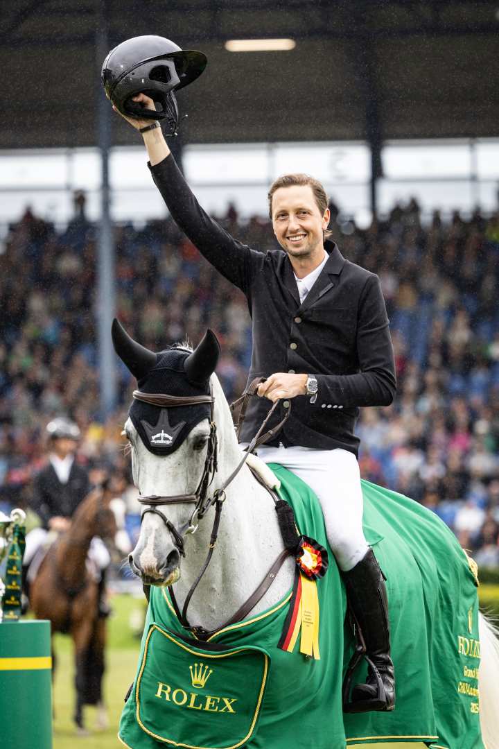 A rider in competition attire sits on a grey horse wearing a green winner's saddlecloth with the Rolex logo. The rider holds his riding helmet aloft in celebration. In the background, another horse with a rider and a packed grandstand in the stadium can be seen.