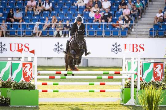 A rider jumps with his horse over a coloured obstacle at a show jumping competition in the stadium. In the background, spectators sit on blue grandstand seats and advertising banners with UBS logos are visible.