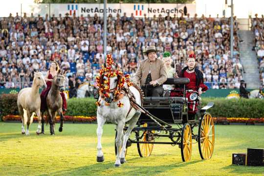 A grey horse decorated with yellow and white flowers pulls a carriage through the main stadium at the opening ceremony. Behind it is a rider with another horse performing a liberty dressage routine.