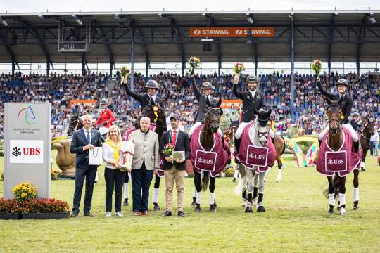 Award ceremony at the CHIO Aachen on the grass of the main stadium. Four riders sit on their horses in competition dress, wearing winning blankets with the UBS logo and holding up bouquets of flowers. Several people, including officials and guests of honor, stand in front of the horses with gifts and flowers. In the background, full grandstands with numerous spectators can be seen, on the left is a stele with the CHIO Aachen and UBS logo.