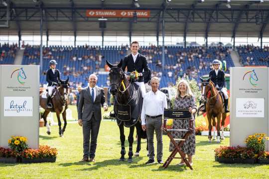 Siegerfoto einer Springreitveranstaltung im Stadion: In der Mitte sitzt der Gewinner in Reitkleidung auf einem schwarzen Pferd, umgeben von drei stehenden Personen. Das Pferd trägt eine Siegerschabracke mit der Aufschrift „Deutschlands U25 Springpokal“. Rechts hält eine Frau einen Regiestuhl mit derselben Aufschrift. Im Hintergrund stehen zwei weitere Reiterinnen auf ihren Pferden. Links und rechts im Vordergrund befinden sich große Tafeln mit Logos des CHIO Aachen sowie Sponsoren, geschmückt mit Blumen. Im Hintergrund sind zahlreiche Zuschauer auf den Tribünen zu sehen.