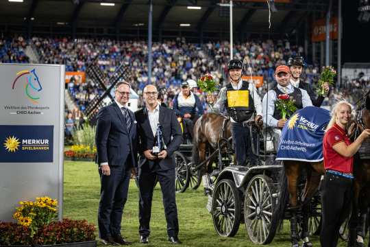 Mehrere Gespannfahrer stehen mit ihren Pferden und Kutschen auf dem Rasen eines Stadions. Sie tragen Siegerdecken mit dem Logo von Merkur Spielbanken und halten Blumensträuße in der Hand. Links im Bild steht eine große Tafel mit dem Logo des CHIO Aachen und von Merkur Spielbanken. Im Hintergrund sind zahlreiche Zuschauer auf den Tribünen zu sehen.