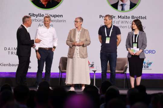Five people, including Michael Mronz, the mayor of Aachen Sibylle Keupen, Prof. Dr. Malte Brettel and Robert Esser, are standing on a stage in front of a large screen displaying names and company logos, apparently at a panel discussion or award ceremony. The audience can be seen in the foreground.