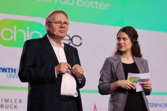 Dressed in a suit, Jörg Howe, Head of Global Communications & External Affairs at Daimler Truck AG, stands next to a woman in a blazer on a stage in front of a screen displaying logos and lettering. The woman is holding presentation cards in her hand.