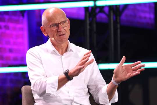 Dr. Dirk Stenkamp, wearing a white shirt, sits on a chair and gestures with his hands. In the background, purple and turquoise streaks of light can be seen, as well as a brick wall. He is sitting on a stage.