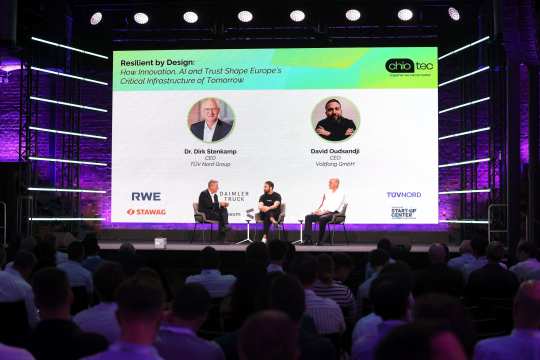 Three people are sitting on a stage discussing in front of a large audience. In the background is a large screen with the title ‘Resilient by Design: How Innovation, AI and Trust Shape Europe's Critical Infrastructure of Tomorrow’ as well as the names, photos and logos of the participating companies. The atmosphere is professional and characterised by modern lighting. The audience can be seen in the foreground.