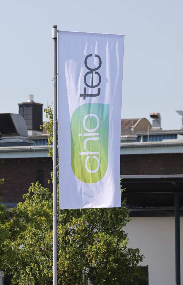 A white flag with the logo and lettering ‘chio tec’ is flying on a flagpole in front of a building and trees under a clear sky.