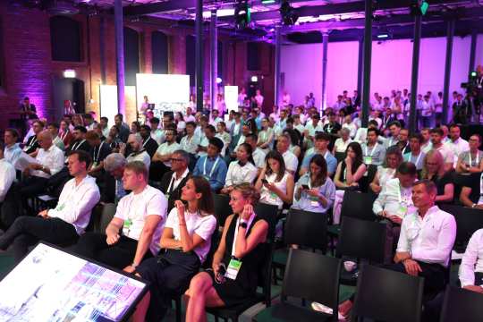 Many people are seated in several rows of chairs in a modern event room with brick walls and purple lighting. Most of the participants are wearing business attire and name tags, and some are standing in the background.
