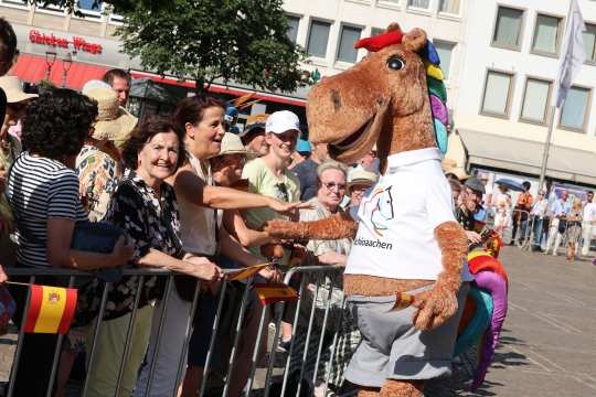 Karli, the CHIO Aachen mascot, a large brown horse on two legs with a colourful mane and a white T-shirt, cheerfully greets people at the Aachen market. Many spectators hold small Spanish flags in their hands.