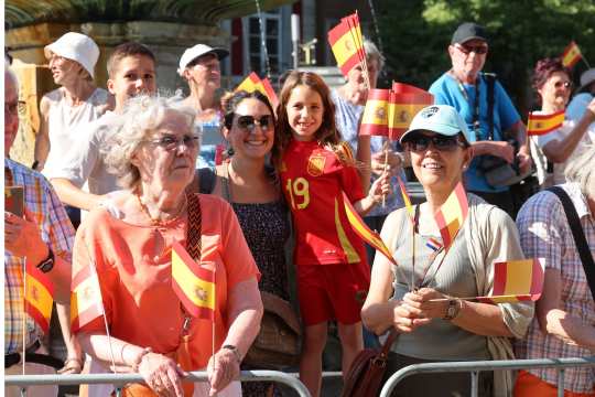 A group of people are standing behind a barrier, holding several Spanish flags. In the middle, a girl is wearing a red Spain jersey with the number 19 on it. The atmosphere is cheerful and the scene is set in sunny weather on the market square in Aachen.