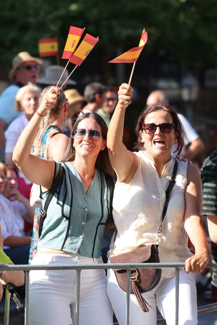 Zwei Frauen stehen hinter einer Absperrung und schwenken mehrere spanische Flaggen in die Luft. Sie tragen sommerliche Kleidung und wirken fröhlich. Im Hintergrund sind weitere Menschen und spanische Flaggen zu sehen. Die Szene spielt sich bei sonnigem Wetter auf dem Aachener Markt ab.