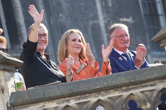 Her Royal Highness Elena of Borbón, Infanta of Spain, the Vice-President of the Aachen-Laurensberger Rennverein, Frank Kemperman, and Aachen's Mayor Sibylle Keupen stand on the balcony of the town hall in front of Aachen's market square, waving and applauding the crowd at a festive event. The historic façade of the town hall can be seen in the background.