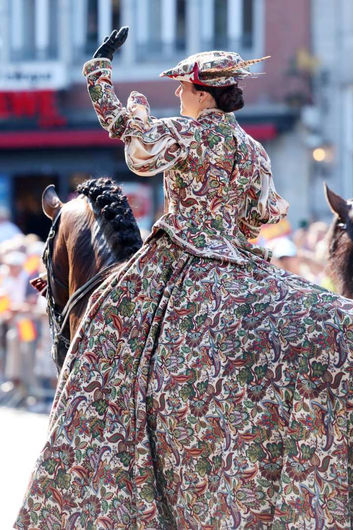 A horsewoman in an elaborately patterned, historical dress with a matching hat sits on a brown horse with a braided mane and waves to the crowd. Spectators and buildings in Aachen city centre can be seen in the background.