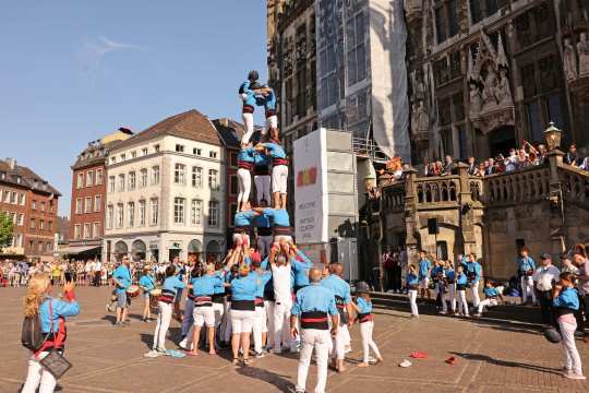 On the bustling market square in front of Aachen's historic town hall, numerous people in blue shirts and white trousers are building a multi-storey human pyramid. Bystanders secure the pyramid while spectators and musicians watch the proceedings.
