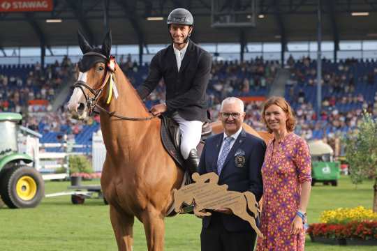 A rider sits on a brown horse with a bow on its bridle on the grass of a show jumping arena. Next to the horse are a man in a suit holding a wooden silhouette of a show jumping horse and a woman in a patterned dress. Spectator stands, flowerbeds and a green vehicle can be seen in the background.