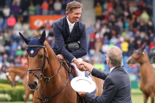 A rider in a dark blue jacket sits on a brown horse with a blue bowler hat and shakes the hand of an official on the showground, who presents him with a silver award. Other horses and spectators in the stands can be seen in the background.