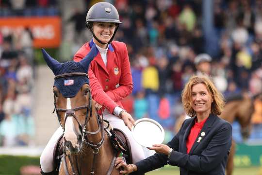 A rider in a red jacket and white breeches sits on a brown horse with a blue fly hat and accepts a silver award from an official on the showground. Spectators in the stands can be seen in the background.