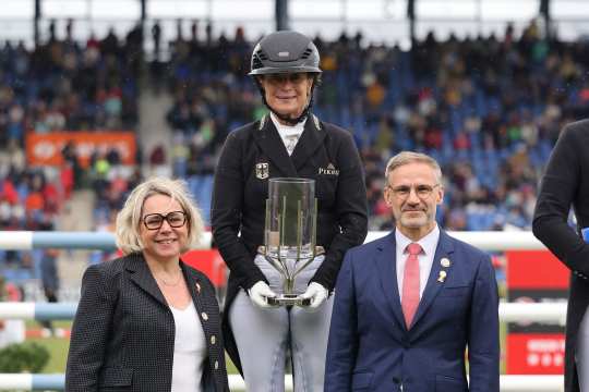 A female rider in a black competition jacket and helmet stands on the podium holding a large glass trophy in her hands. Next to her are a woman in a blazer and a man in a blue suit with a red tie. Spectators can be seen in the stands in the background.