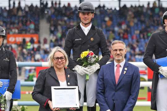 A rider in black competition dress and helmet stands on the podium holding a bouquet of flowers in his hands. Next to him are a woman with a certificate and a man in a blue suit with a red tie. Spectators can be seen in the stands in the background.